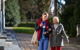 Two women walking outdoors on a path with trees and grass in the background

