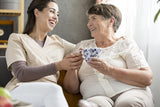 Two women sitting together, one holding a cup, in a warm indoor setting.

