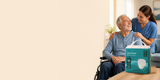 Smiling older man in a wheelchair with a carer, with a pack of Live N Care Adult Pull-Up Pants on the table